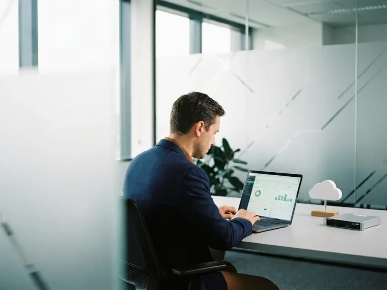 Business professional analyzing cloud cost optimization dashboard on laptop at modern corporate desk with server model nearby.
