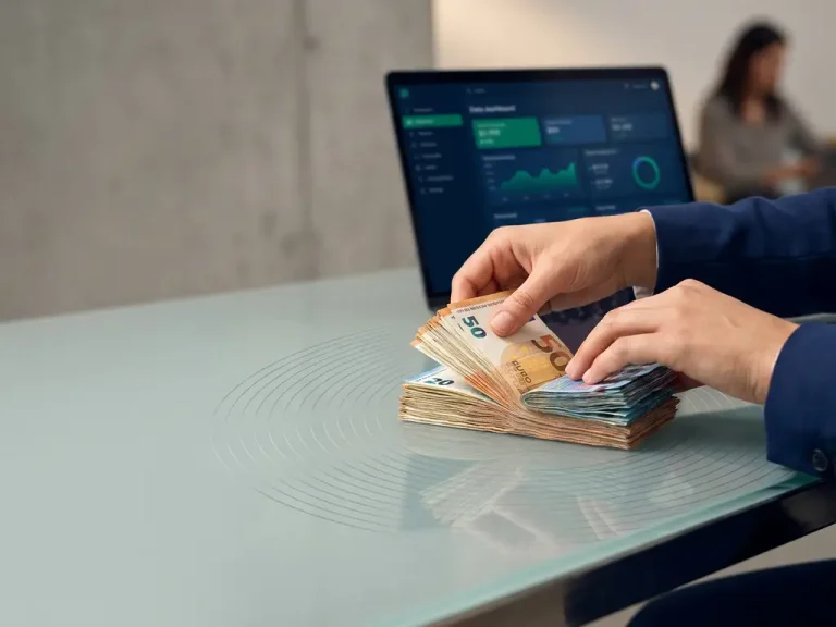 Hands splitting a stack of euro banknotes into two equal portions on a glass desk, with a glowing laptop dashboard in the background.